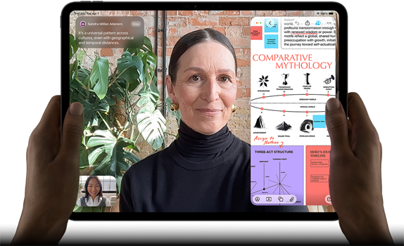 iPad Pro, front exterior, landscape orientation, space black colour, rounded corners, black display bezel, hands holding left and right sides, display showing a FaceTime conversation being live translated, and an educational app showing a presentation of "Comparative Mythology" with text, diagrams and handwritten notes