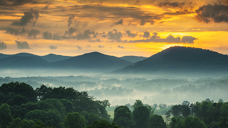 Tree-covered mountains tower above fog-laden woods; the sun is rising Tree-covered mountains tower above fog-laden woods; the sun is rising