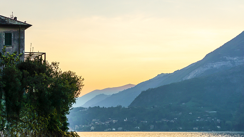 northern Italy - a house overlooks the water in the foreground, with a coastal town at the foot of a mountain in the background northern Italy - a house overlooks the water in the foreground, with a coastal town at the foot of a mountain in the background